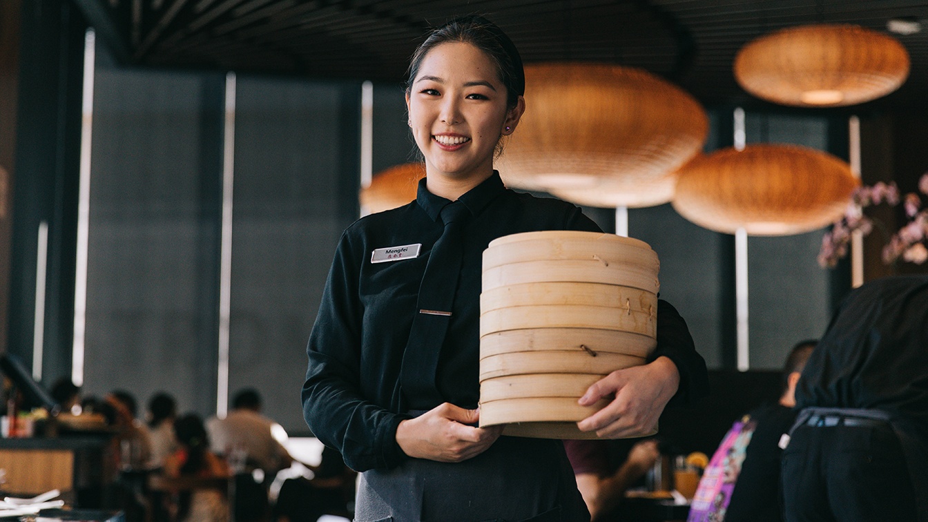 A server smiling and holding a bamboo basket of Xiao Long Baos.